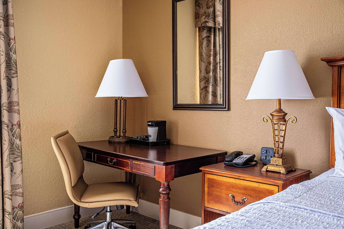 A hotel room with a desk, chair, bedside table, lamp, and a bed.