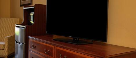 A hotel room with a wooden dresser, a flat-screen TV, a refrigerator, and a patterned armchair.