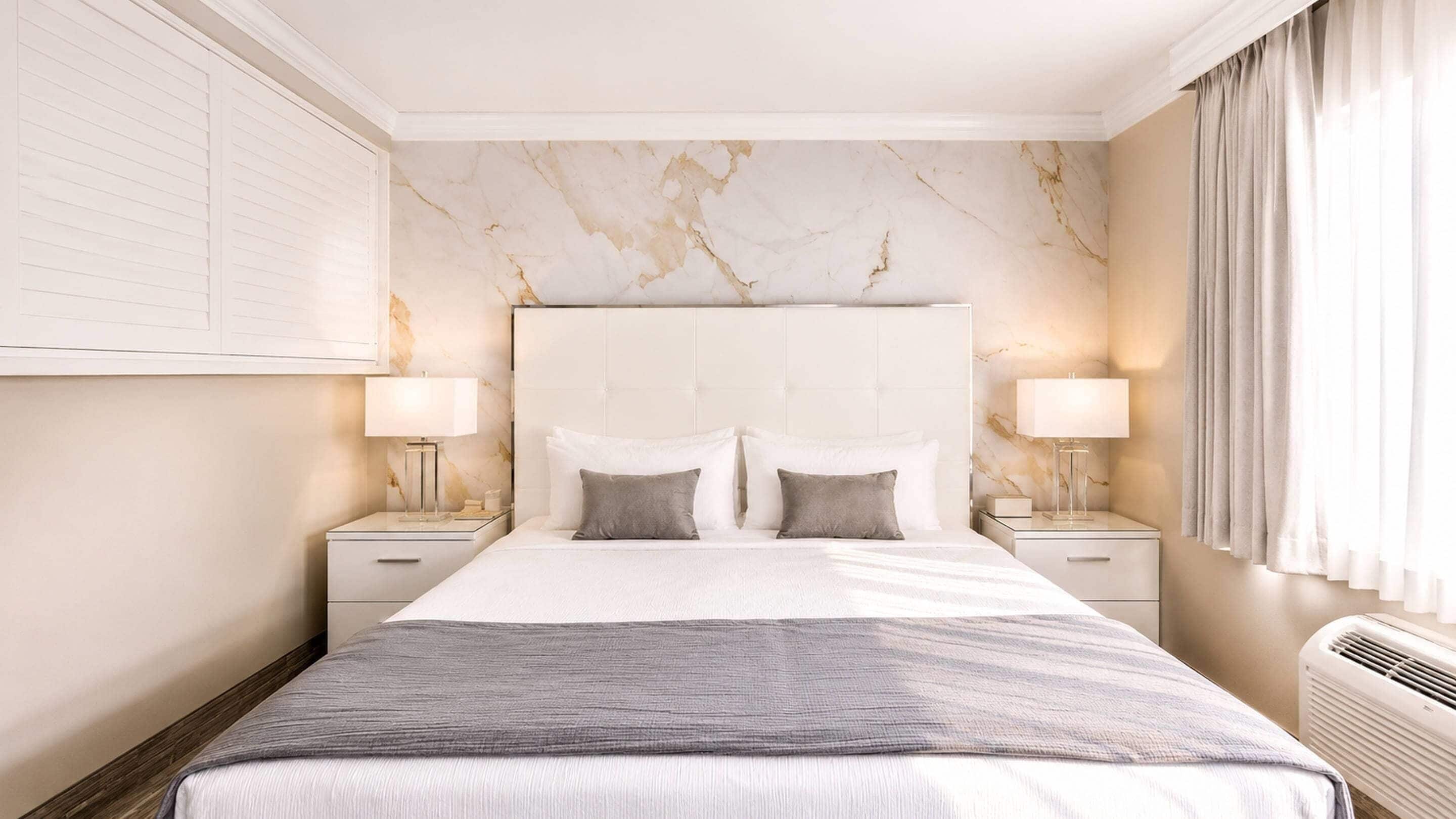 Une chambre d’hôtel avec une tête de lit en marbre, deux tables de chevet avec des lampes, un grand lit recouvert d’une couverture grise et une fenêtre avec des rideaux.