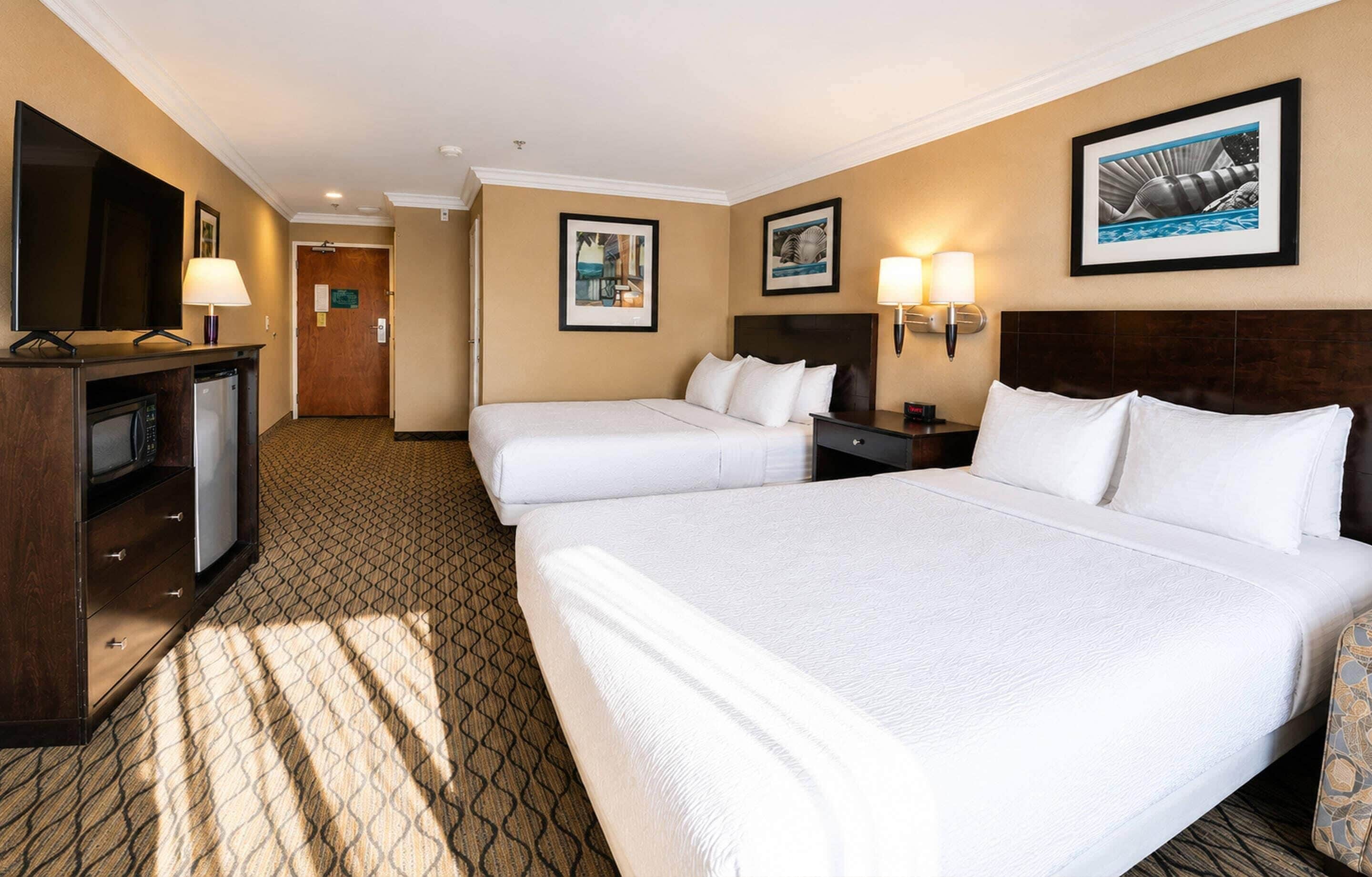 A hotel room with two beds, a desk with a television, and framed pictures on the walls.