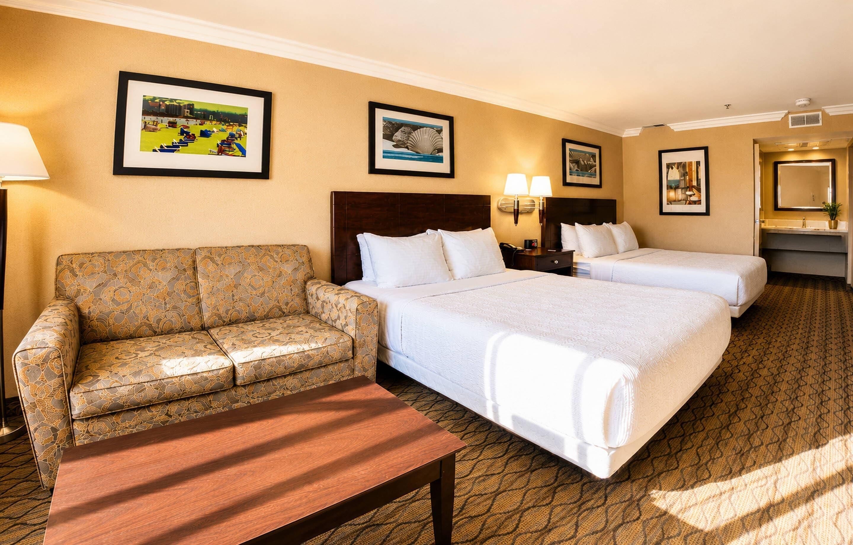 A hotel room with a sofa, two beds, a bedside table, a lamp, and framed pictures on the walls.