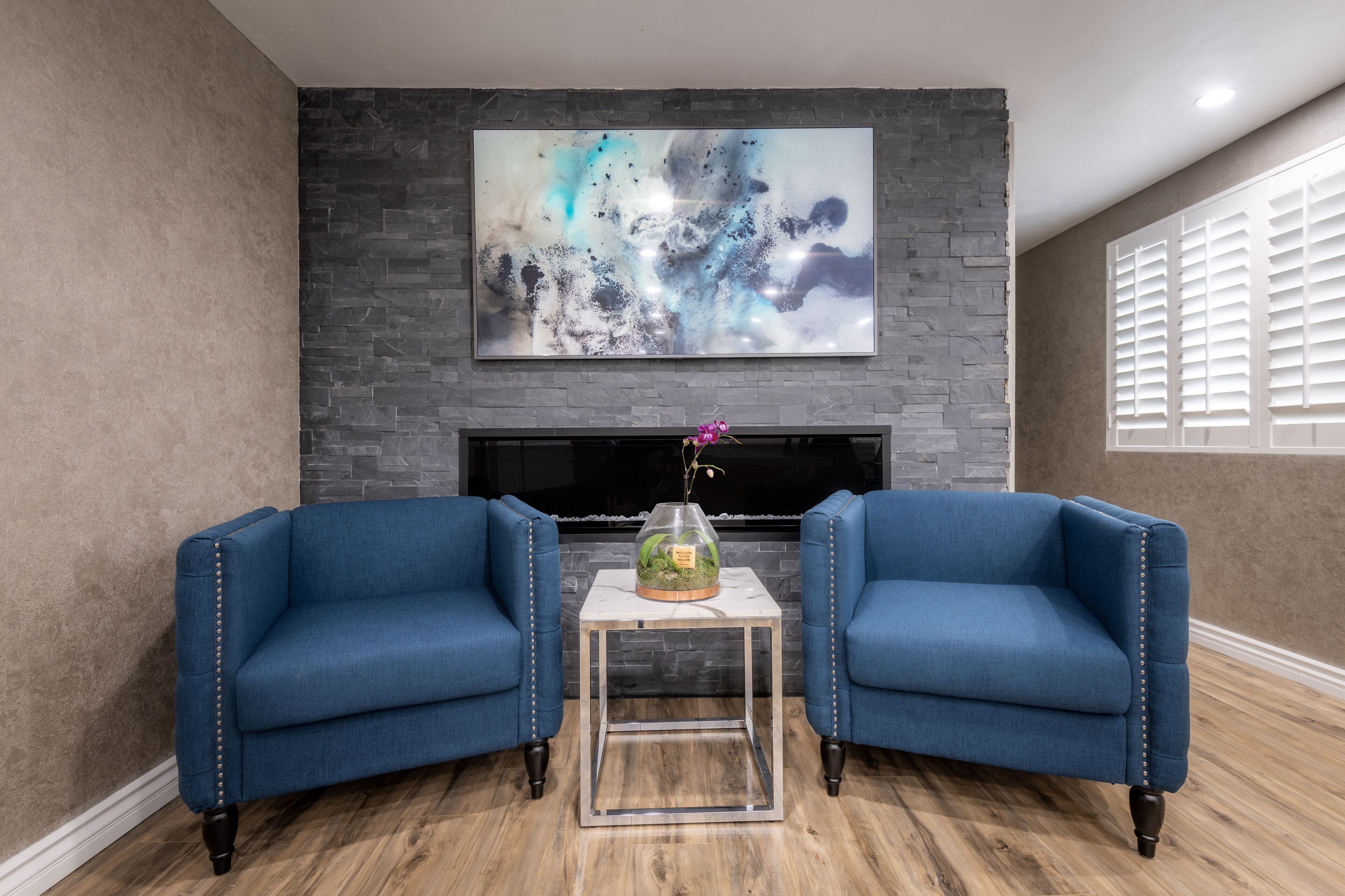 A living room with two blue armchairs, a small table with a vase, and a large abstract painting above a fireplace.