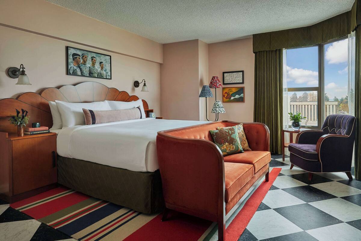 Une chambre d’hôtel avec un grand lit, un canapé, un fauteuil, une fenêtre donnant sur un beau paysage et un tableau au mur.
