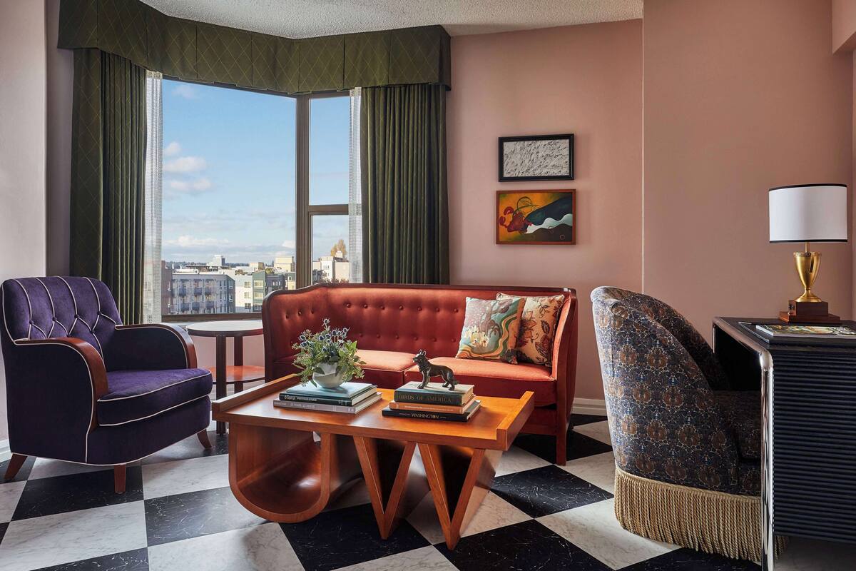 Une chambre d’hôtel avec un canapé rouge, un fauteuil violet, une table basse en bois et une vue sur la ville grâce à de grandes fenêtres.
