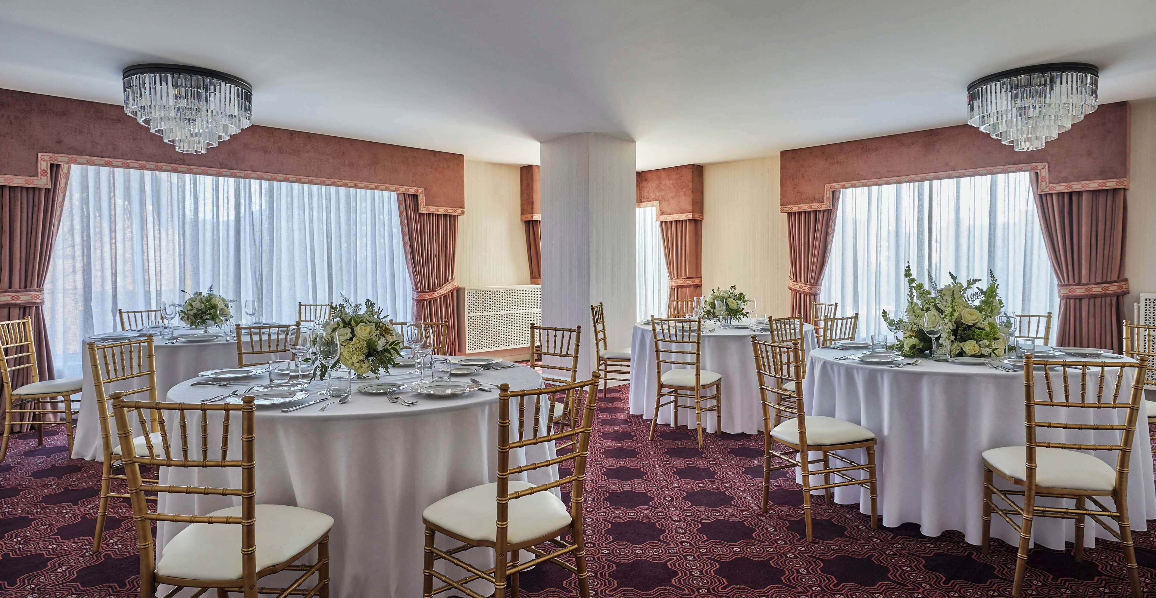A banquet hall with round tables, gold chairs, and chandeliers.