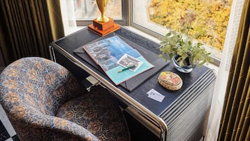 A room with a window view of autumn foliage, a table with a magazine, a lamp, and a small plant.