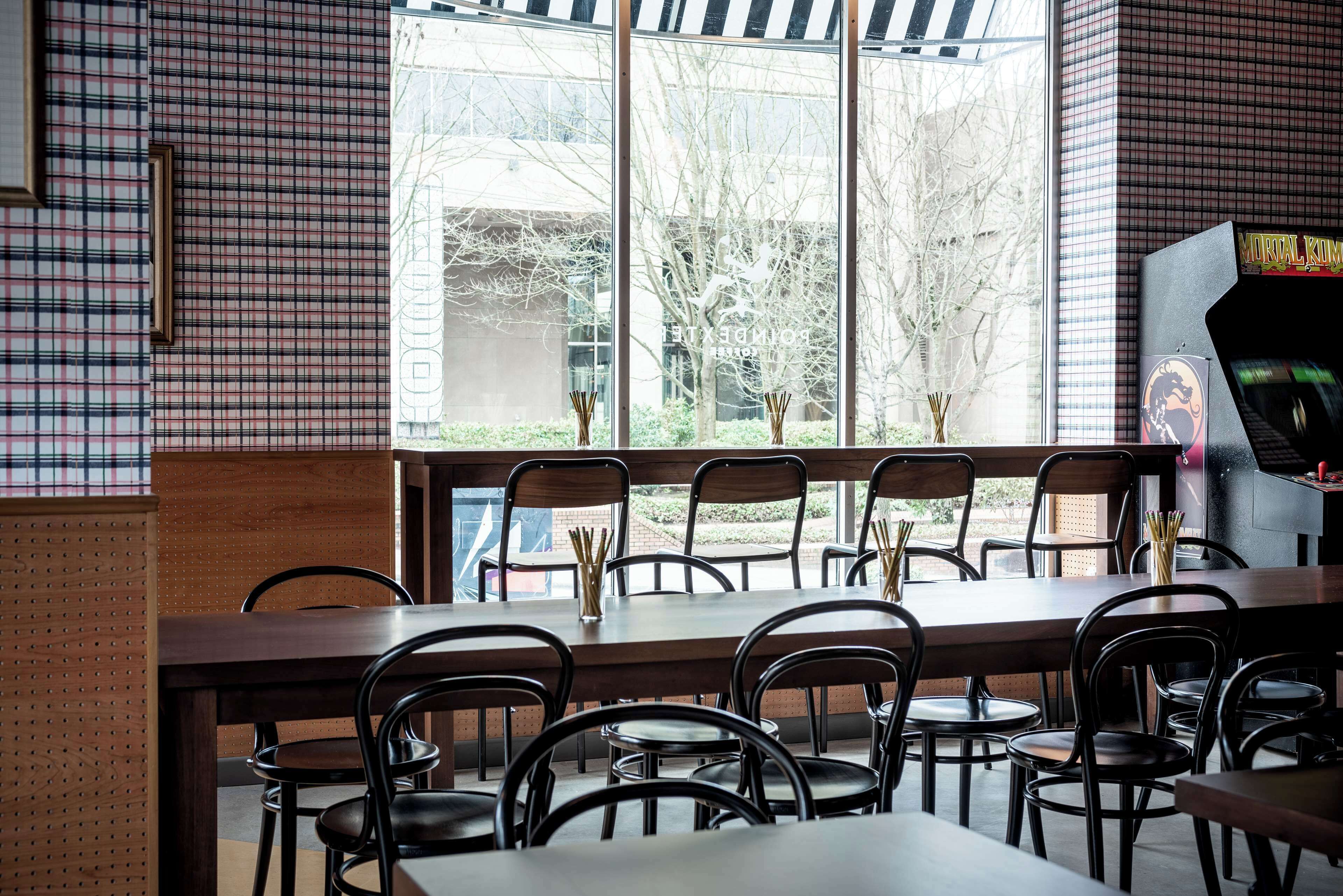 Un restaurant avec une longue table et des chaises, de grandes fenêtres donnant sur des arbres et un bâtiment, et une borne d’arcade située à droite.