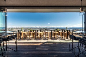 A rooftop terrace with wooden tables and chairs, offering a panoramic view of a city and mountains.