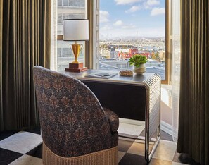 A room with a view of a cityscape, a desk with a lamp, a chair, and a small potted plant.