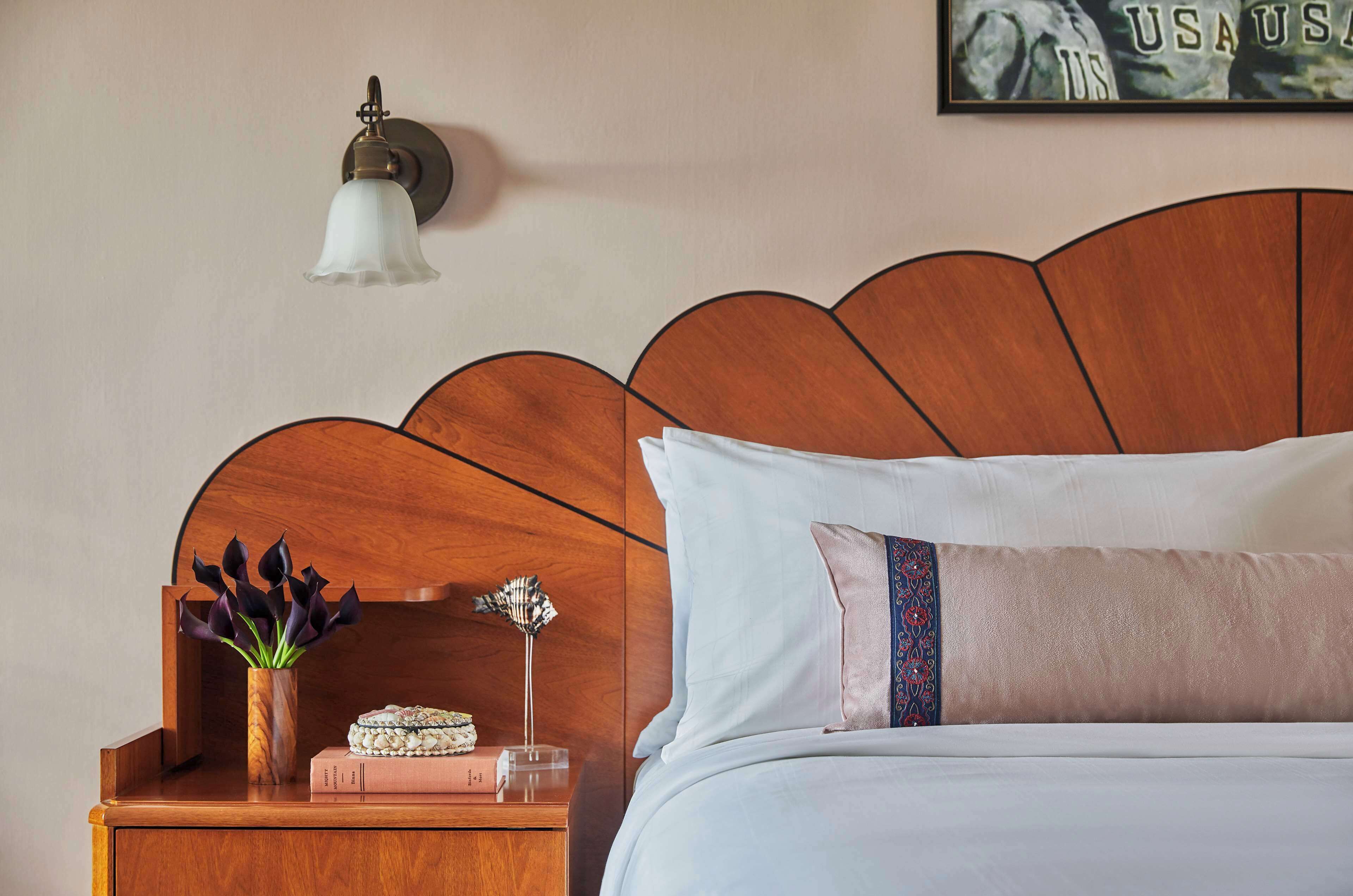 A neatly made bed with a wooden headboard, a bedside table with a vase of flowers, a lamp, and a framed picture on the wall.