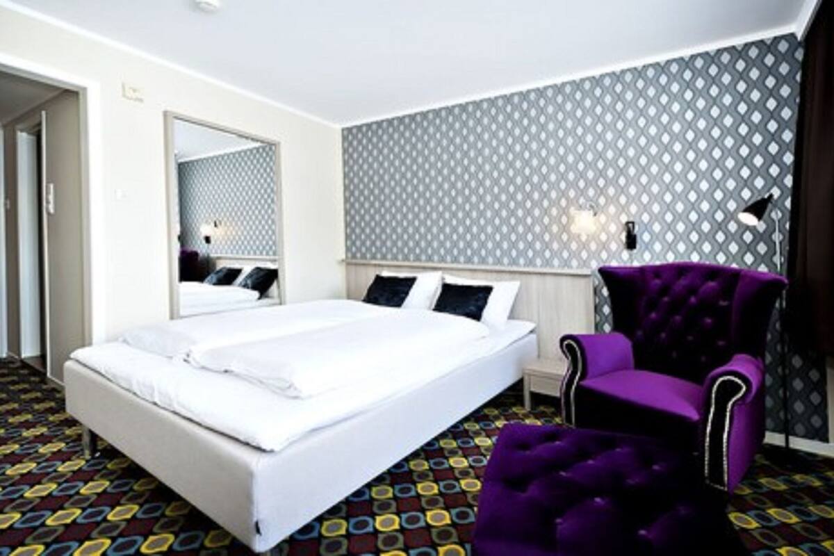 A hotel room with a large bed, a purple armchair, and patterned carpet.