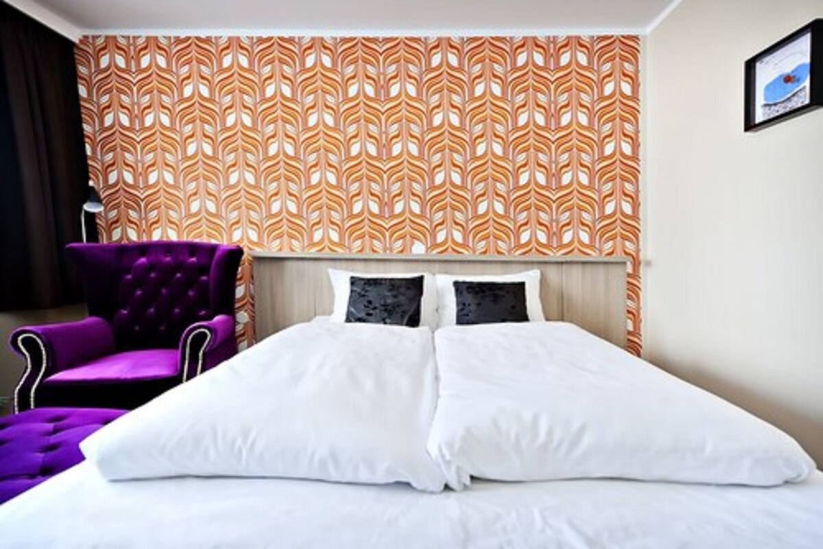 A hotel room with a bed, two pillows, a headboard, a purple armchair, and a patterned wall.