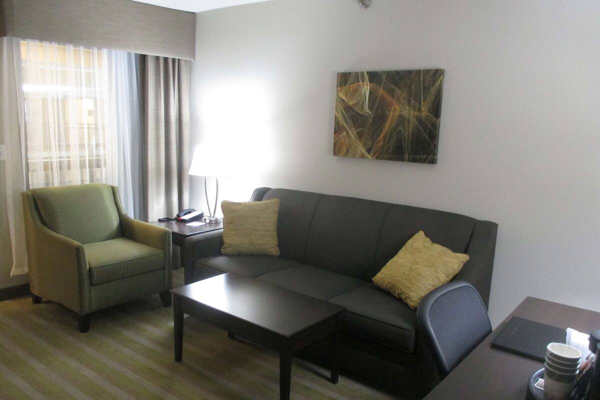 A hotel room with a green armchair, a grey sofa, a coffee table, and a painting on the wall.