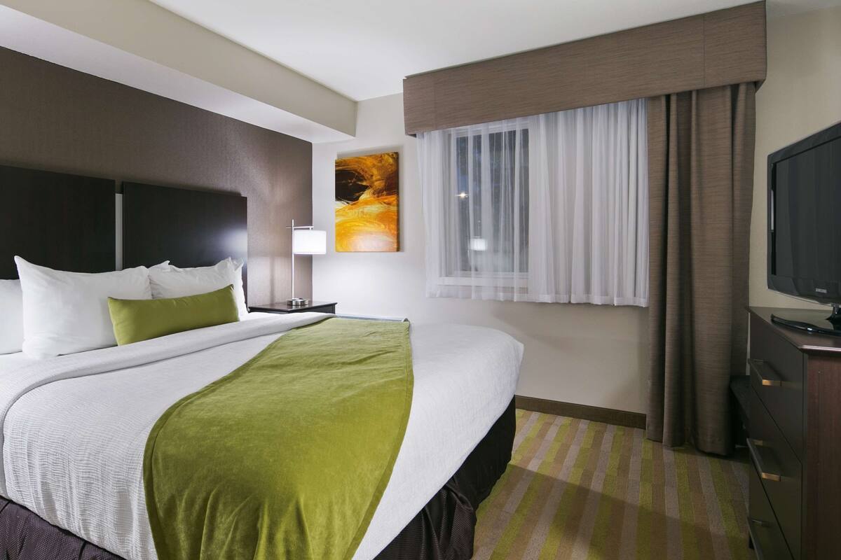 A hotel room with a large bed, a flat-screen TV, a window with curtains, and a painting on the wall.