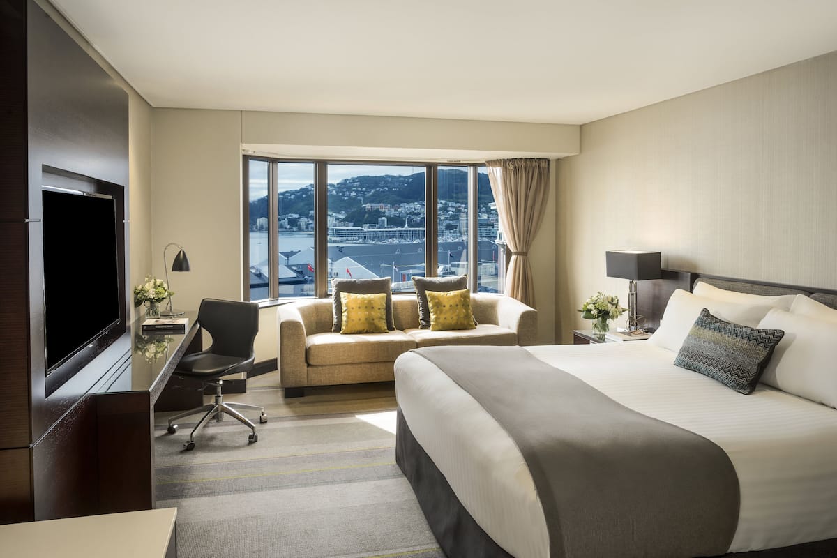 Une chambre d’hôtel avec un grand lit, un bureau avec une chaise, une télévision et une vue sur le paysage urbain grâce à de grandes fenêtres.