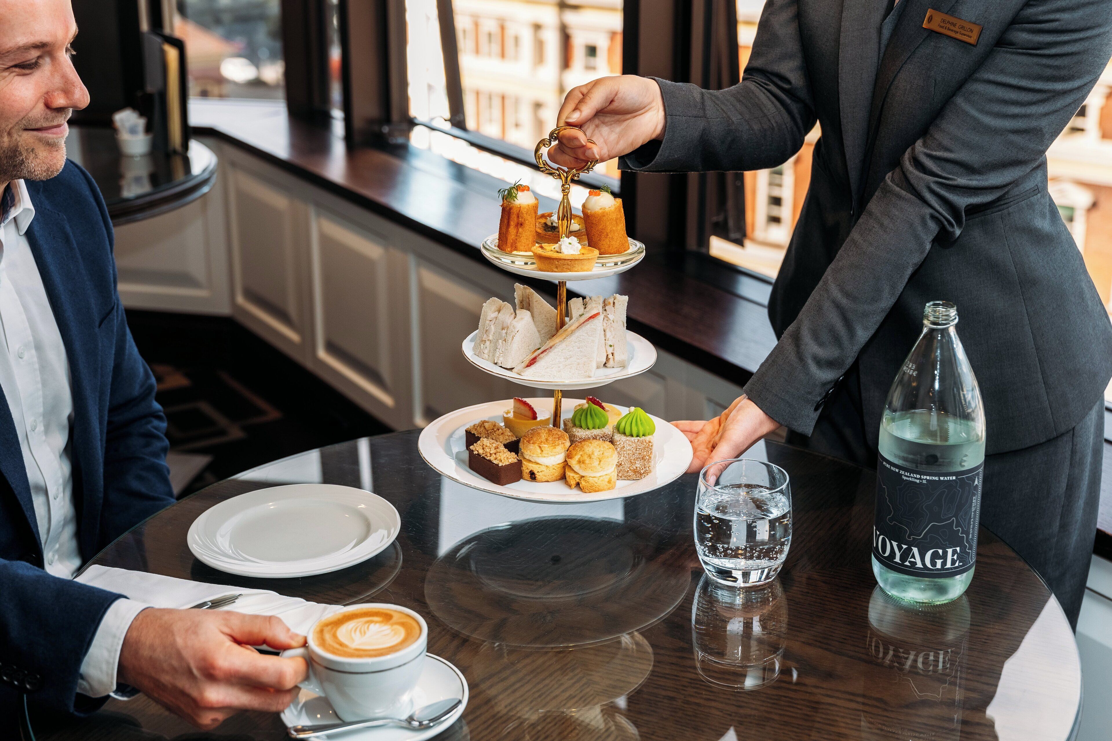 A person serving a multi-tiered dessert tray to a guest at a table with a coffee cup and a bottle of water.