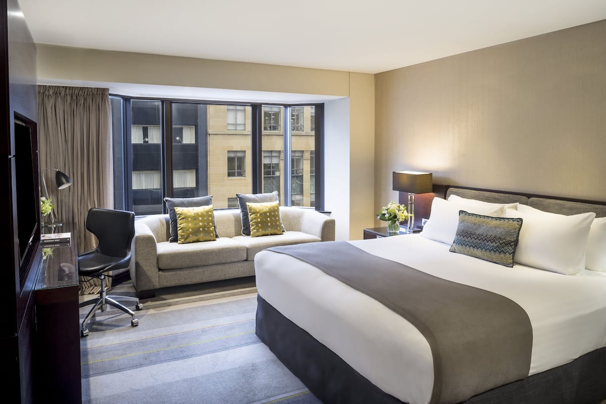 Une chambre d’hôtel avec un grand lit, un canapé, un bureau avec une lampe et une vue sur les immeubles à travers de grandes fenêtres.