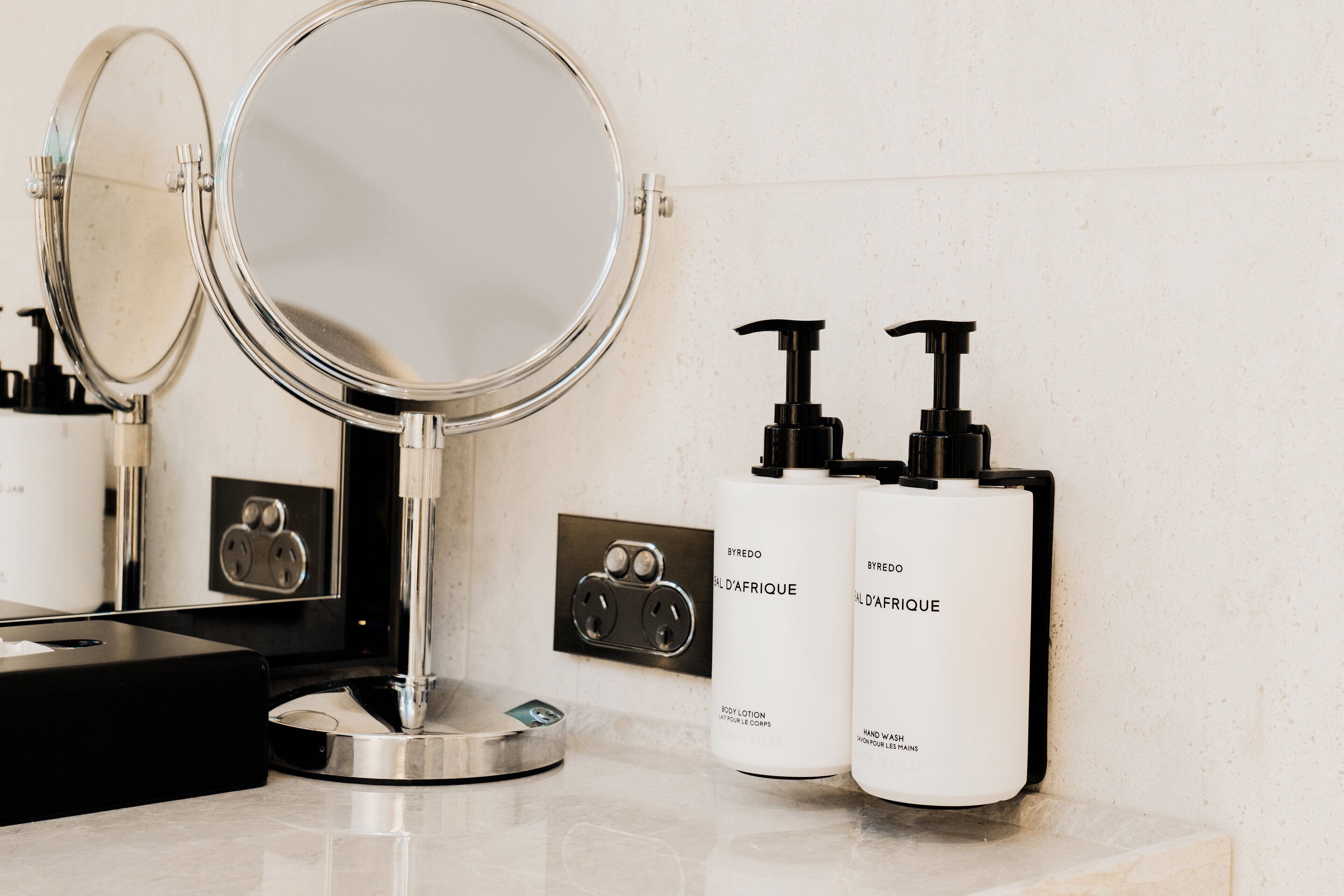 Un tocador de baño con un espejo redondo, dos botellas de crema corporal y un dispensador de jabón negro.