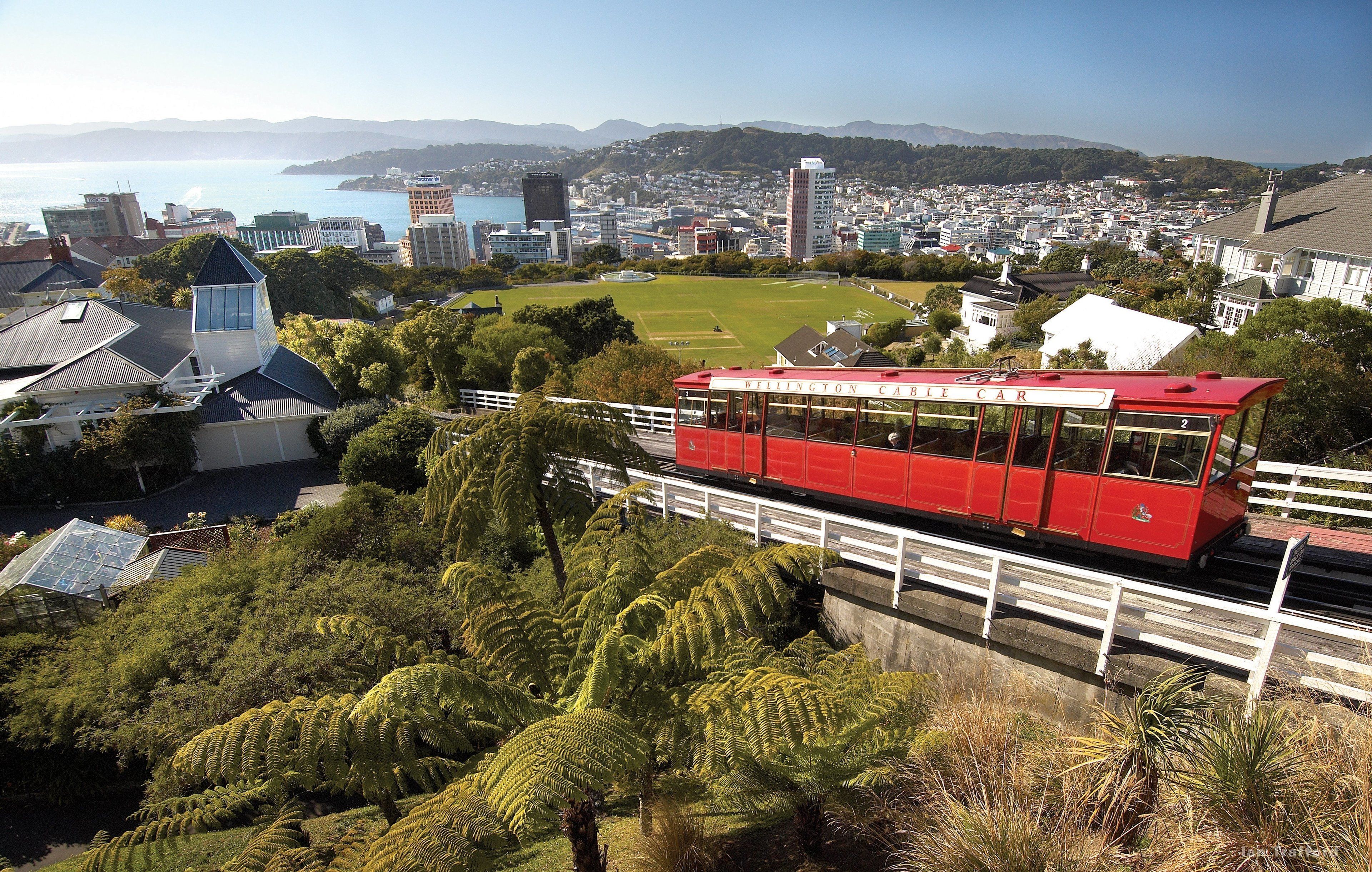 Un funicular rojo con la leyenda 'Wellington Cable Car', que viaja sobre un puente con vegetación y un paisaje urbano al fondo.