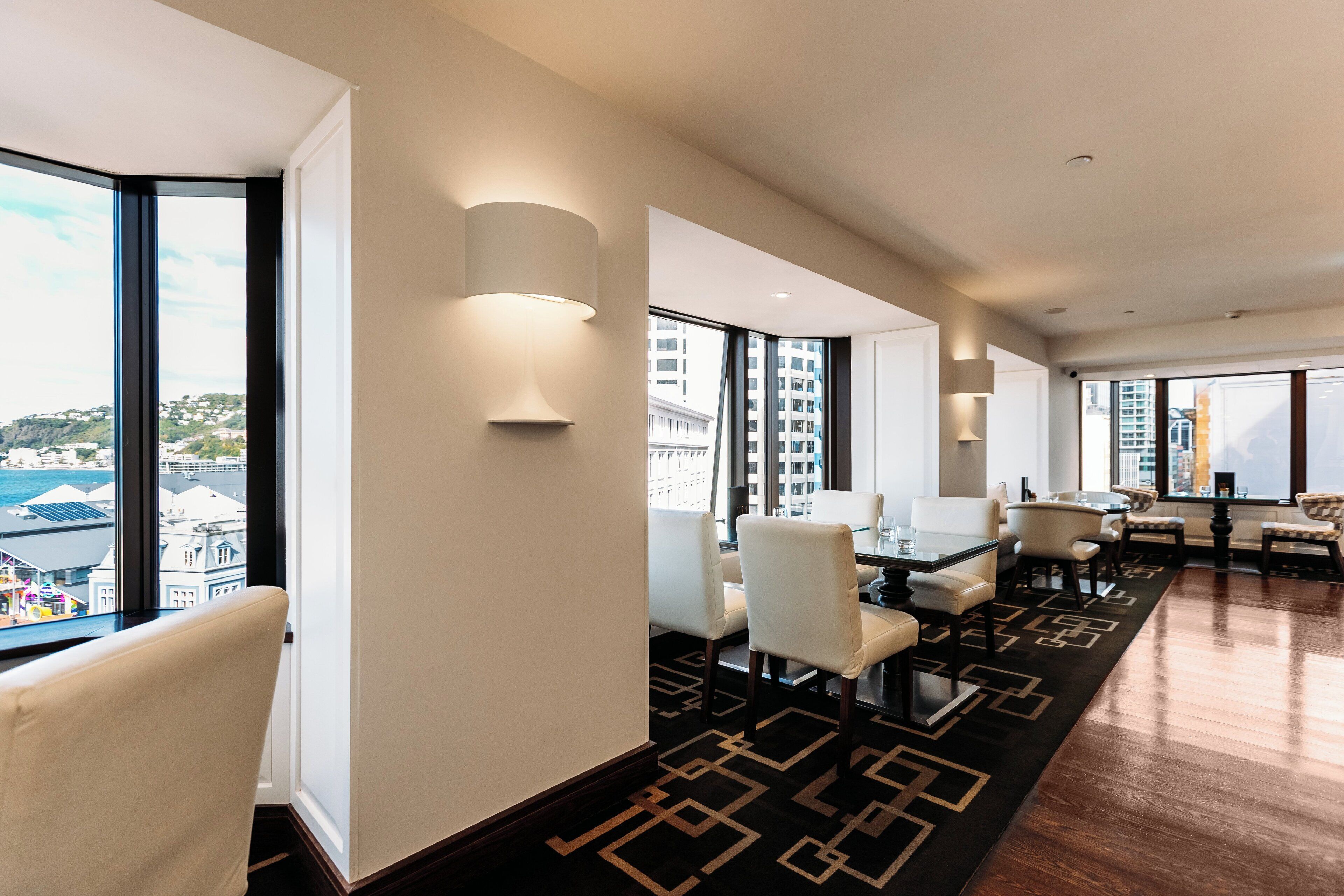 Une chambre d’hôtel moderne dotée d’une grande fenêtre donnant sur la ville, d’un coin repas agrémenté d’un tapis à motifs et d’un espace salon.