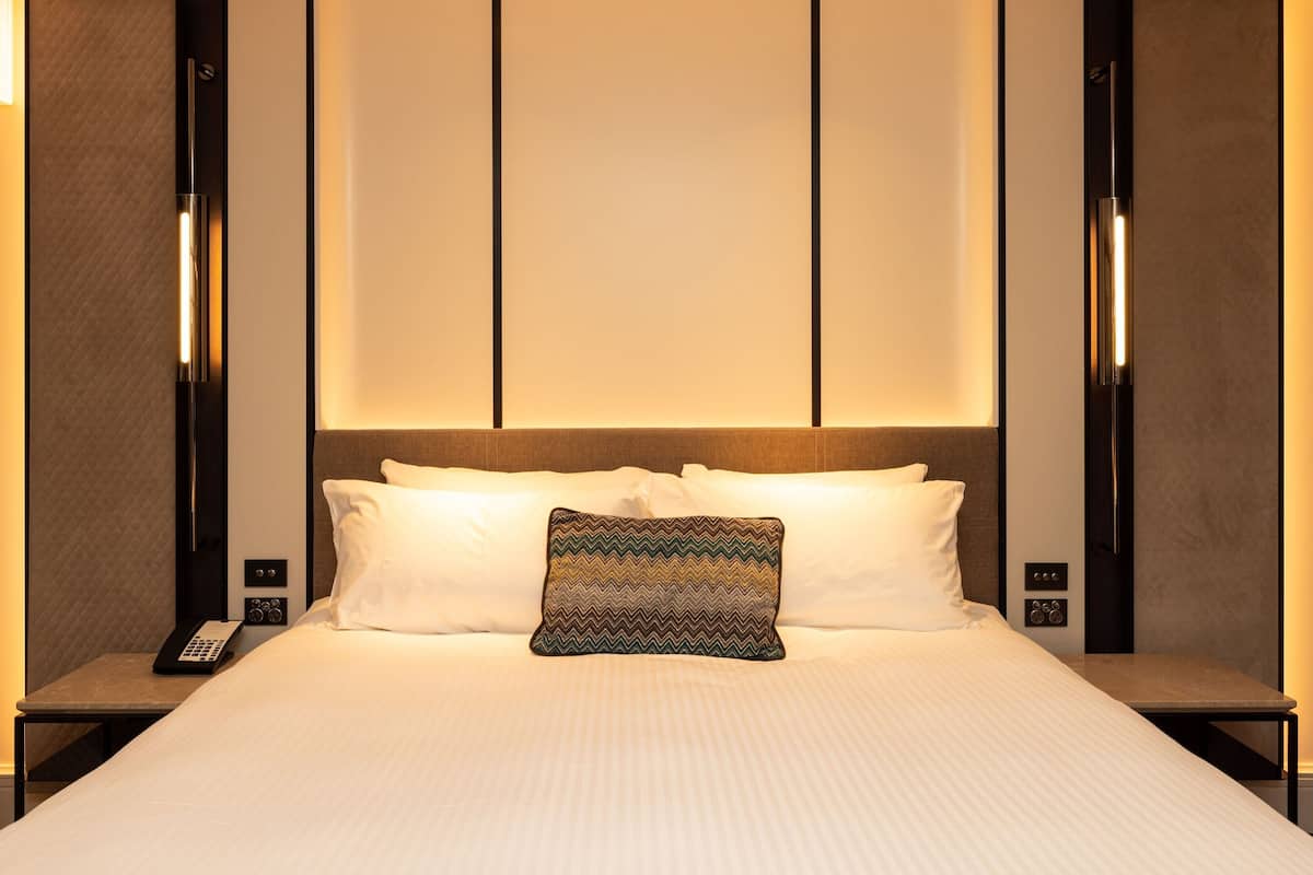 Une chambre d’hôtel avec un grand lit, deux tables de chevet, une petite table d’appoint, un coussin décoratif à motifs et un luminaire fixé au mur.