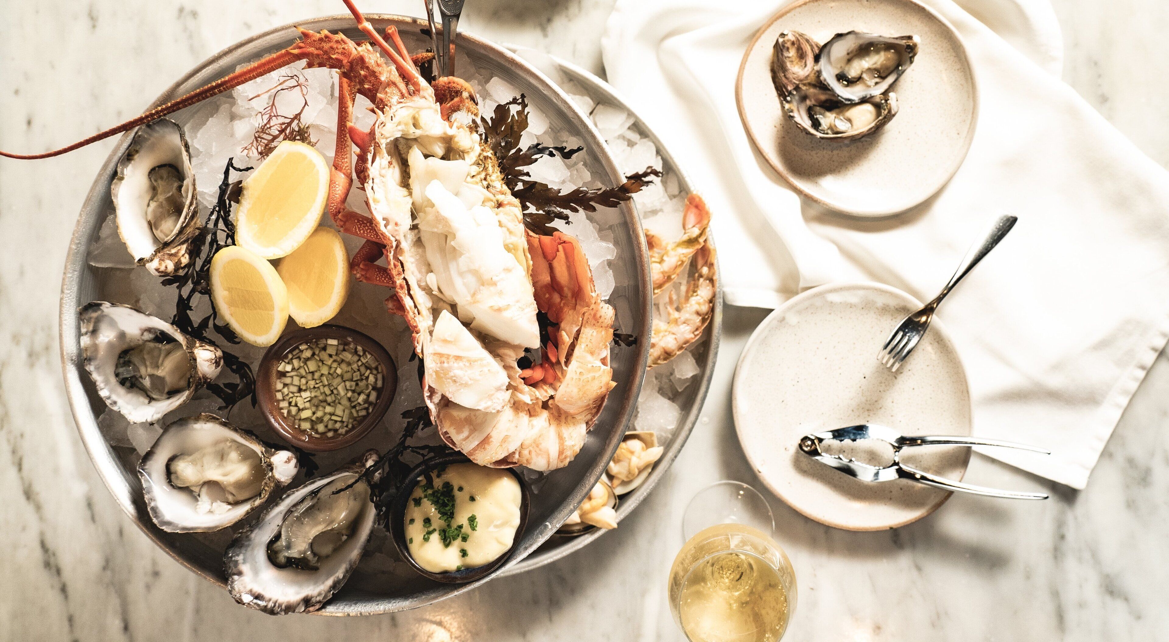 A seafood platter with various shellfish and a bowl of sauce, accompanied by a glass of white wine.