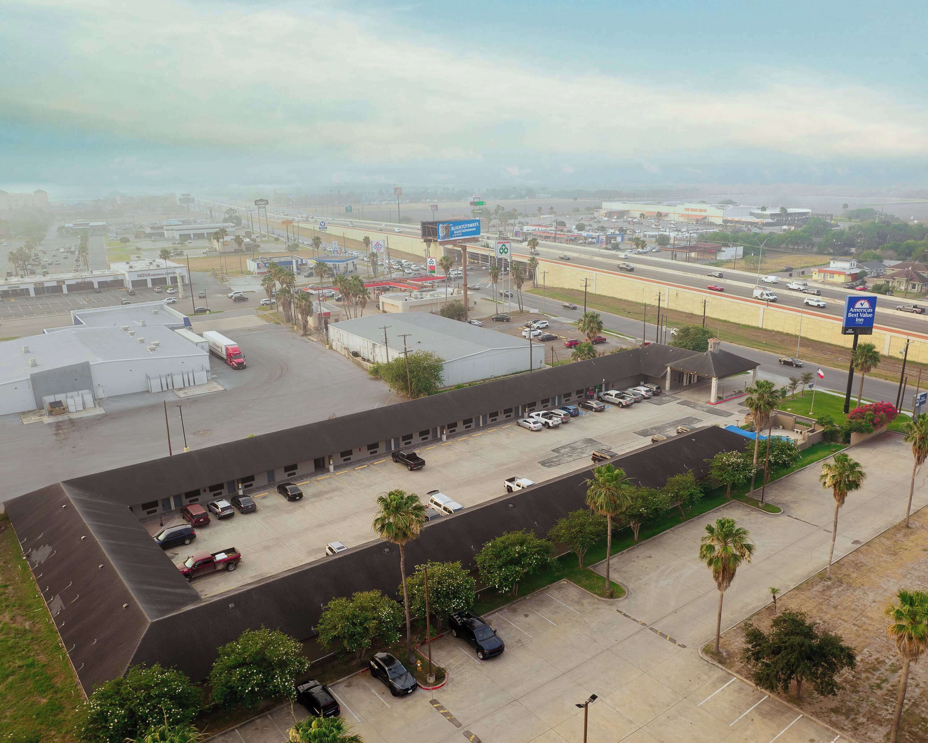 A large parking lot with multiple cars, a building with a flat roof, and a highway in the background.