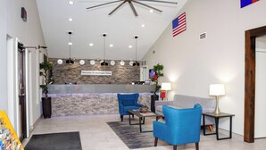 A hotel lobby with a reception desk, two blue armchairs, a grey sofa, a small table with a lamp, and a large American flag.