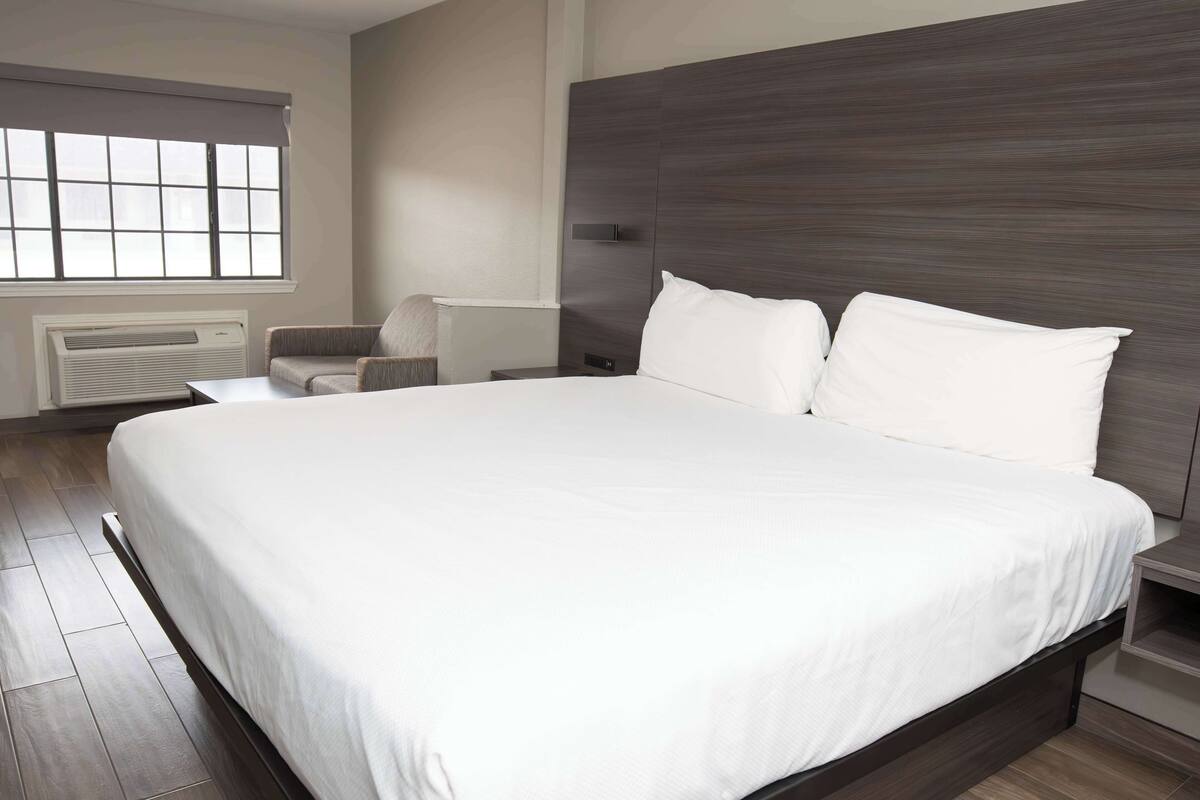 A modern bedroom with a large bed, a chair, a nightstand, and a window with blinds.