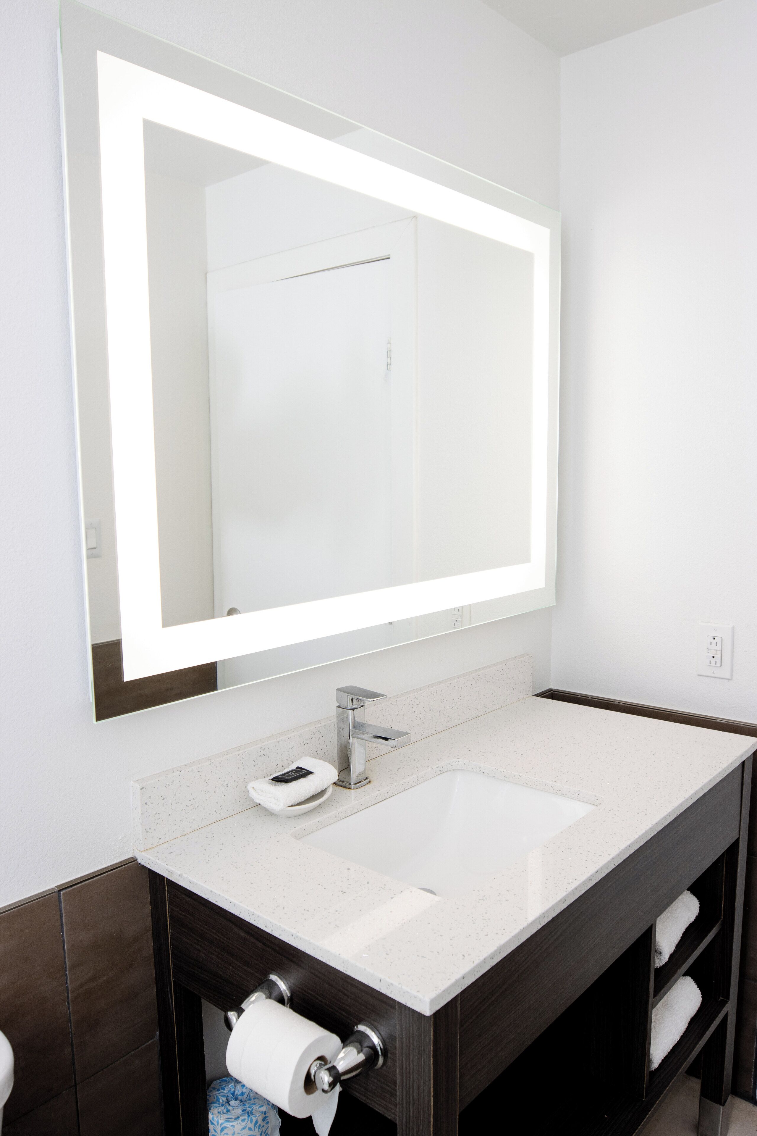 Un baño con un espejo grande, lavamanos blanco, tocador de madera oscura y un perchero para toallas.