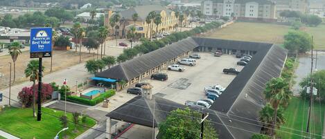 A hotel with a parking lot, a swimming pool, and a large building in the background.