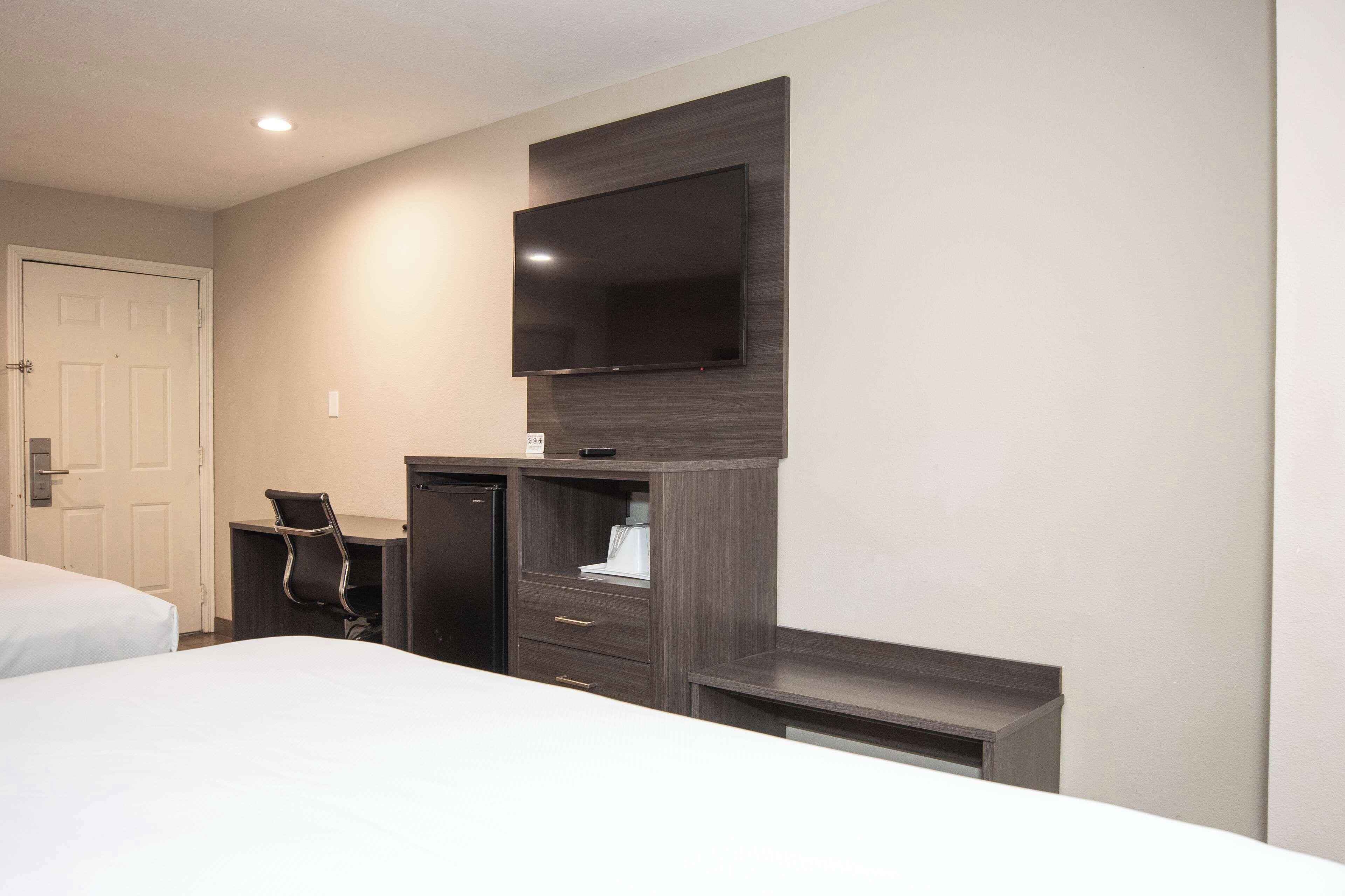 A hotel room with a bed, a desk with a chair, a flat-screen TV mounted on the wall, and a door.