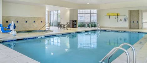 Una piscina interior con agua azul cristalina, rodeada de paredes y piso de azulejos. Se observan escaleras metálicas de piscina, y un conjunto de sillas y mesas cerca de la piscina.