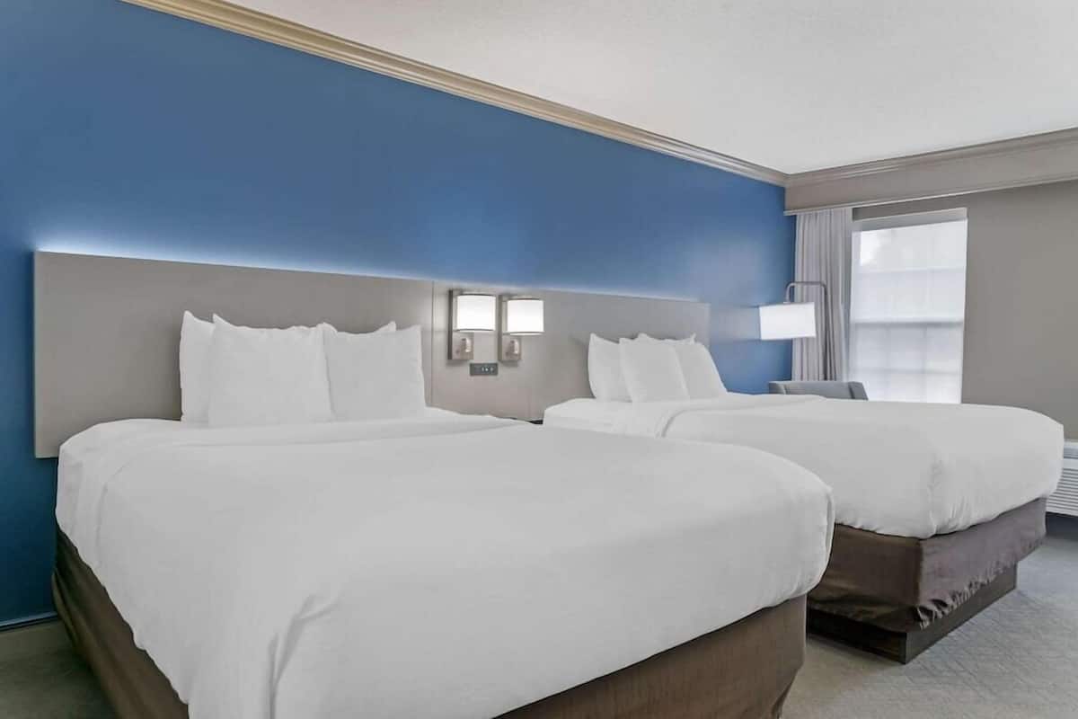A hotel room with two beds, a large headboard, bedside lamps, a window with curtains, and a flat-screen TV.