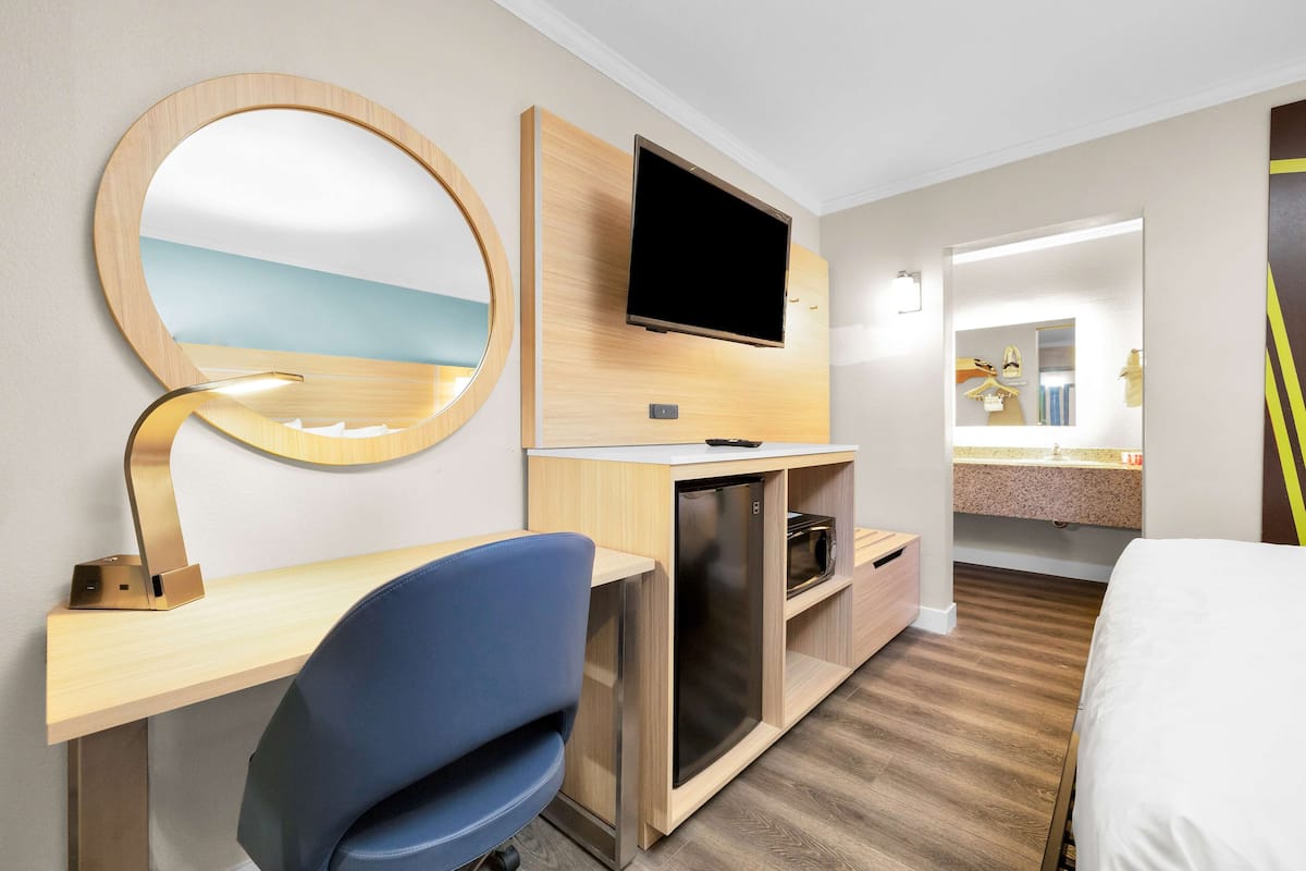 Hotellihuone, jossa on puinen työpöytä, sininen tuoli, televisio, peili ja sänky.