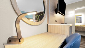 Una habitación de hotel moderna con un escritorio de madera, una silla azul, un espejo redondo, un televisor de pantalla plana montado en la pared y una mesita de noche pequeña.