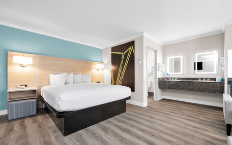 A modern hotel room with a large bed, bedside tables, a built-in bench, a vanity with a mirror, and a shower area.