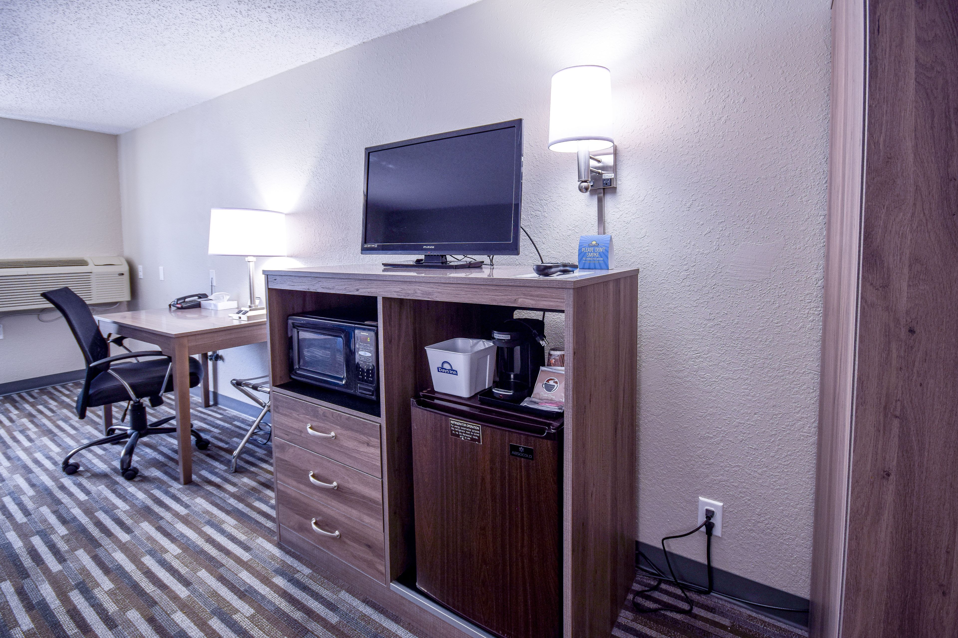 A hotel room with a wooden desk, a computer monitor, a lamp, a microwave, a refrigerator, and a chair.