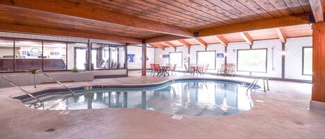 An indoor swimming pool with a wooden ceiling, tiled floor, and several tables and chairs around the pool area.