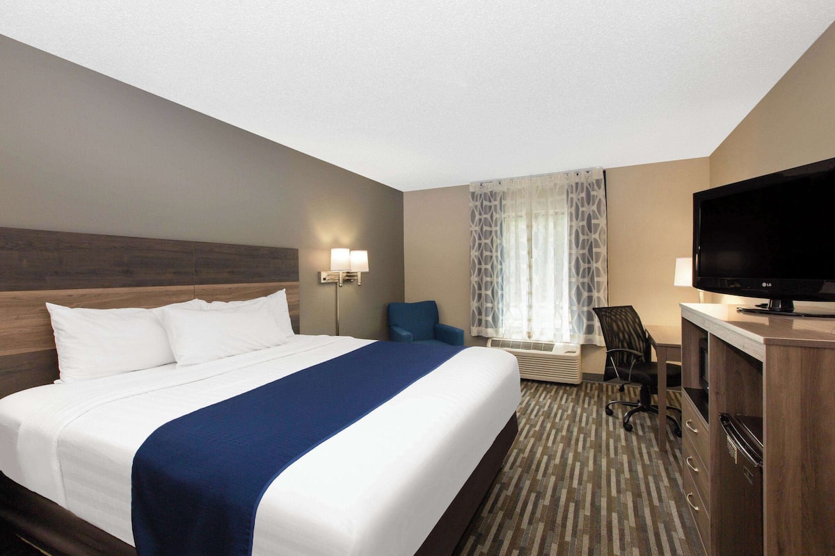 A hotel room with a large bed, a desk with a flat-screen TV, a chair, a lamp, and a window with curtains.
