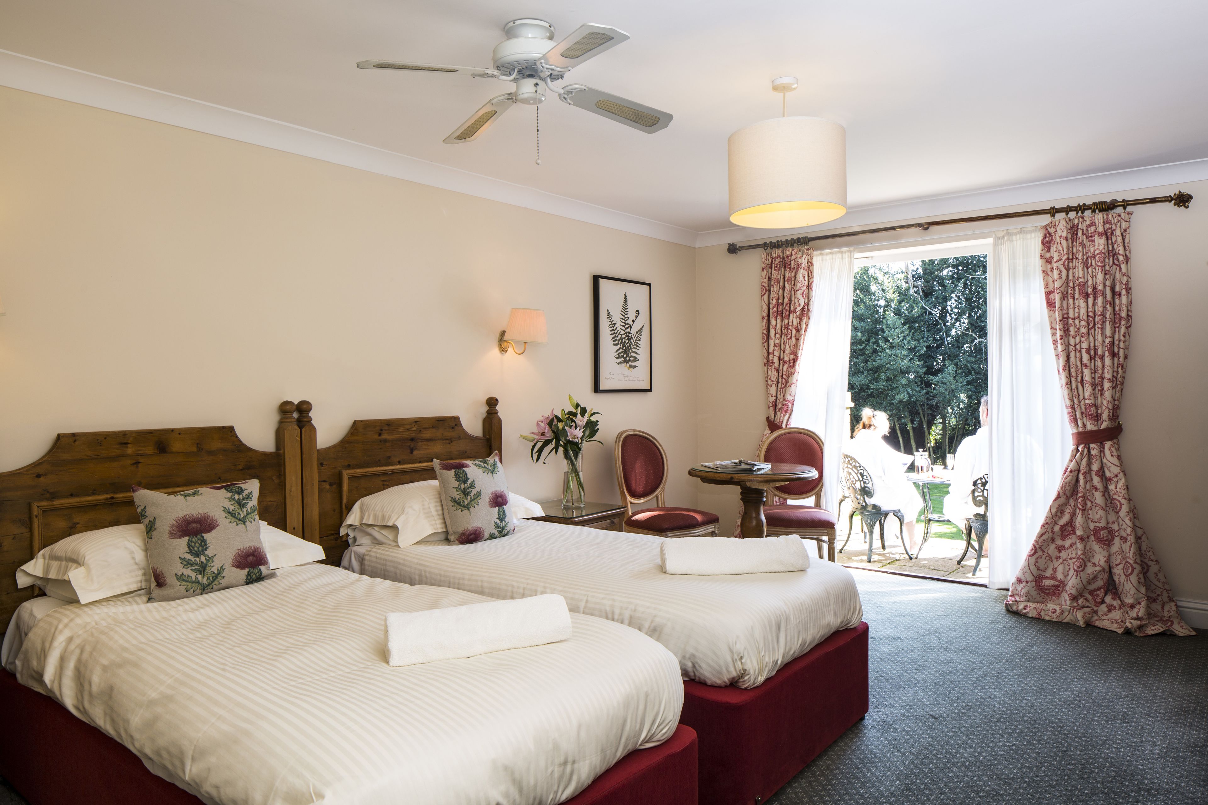 A hotel room with two beds, a ceiling fan, a table with chairs, and a view of the outdoors.