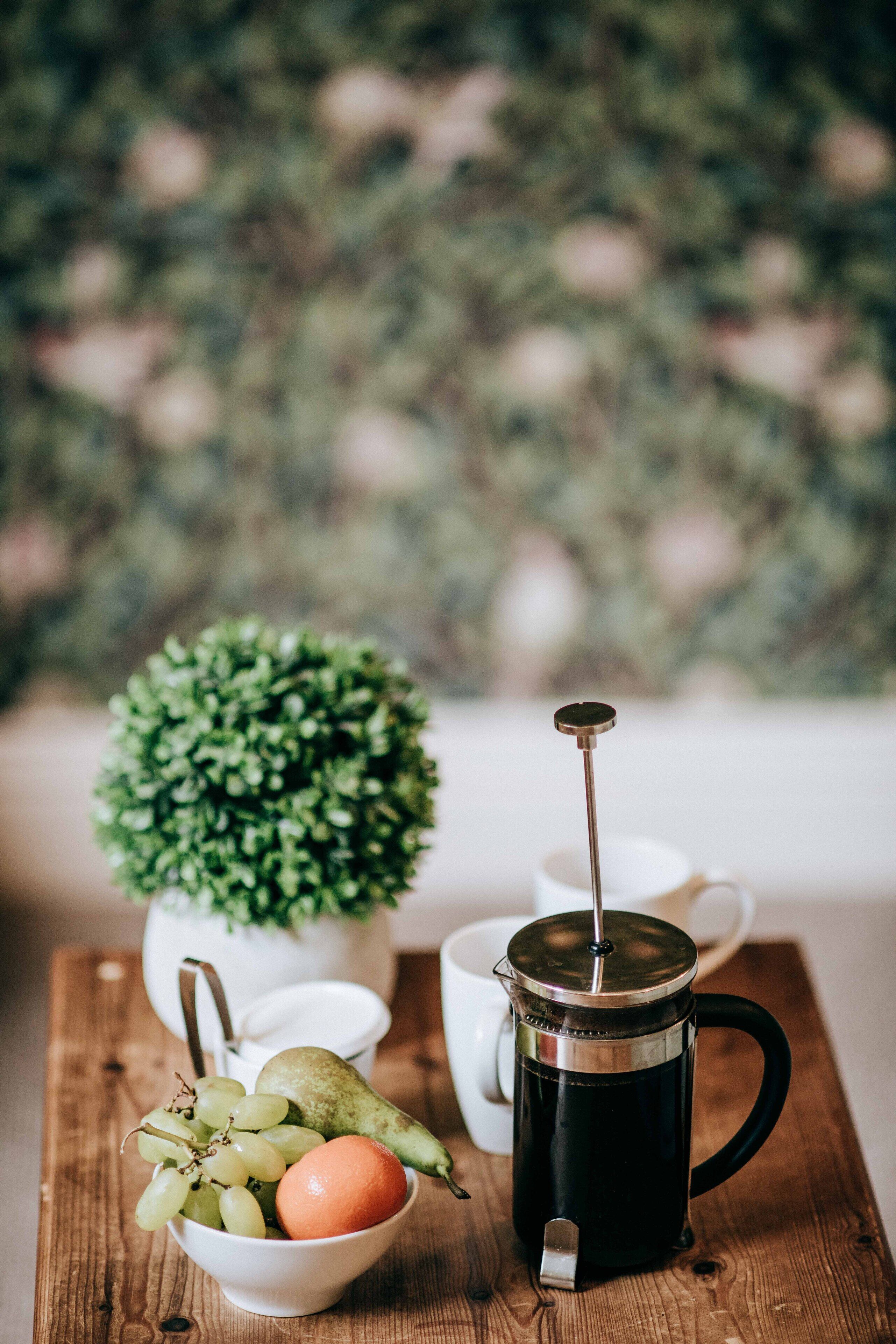 A coffee maker, a bowl of fruit, and a small potted plant on a wooden table.