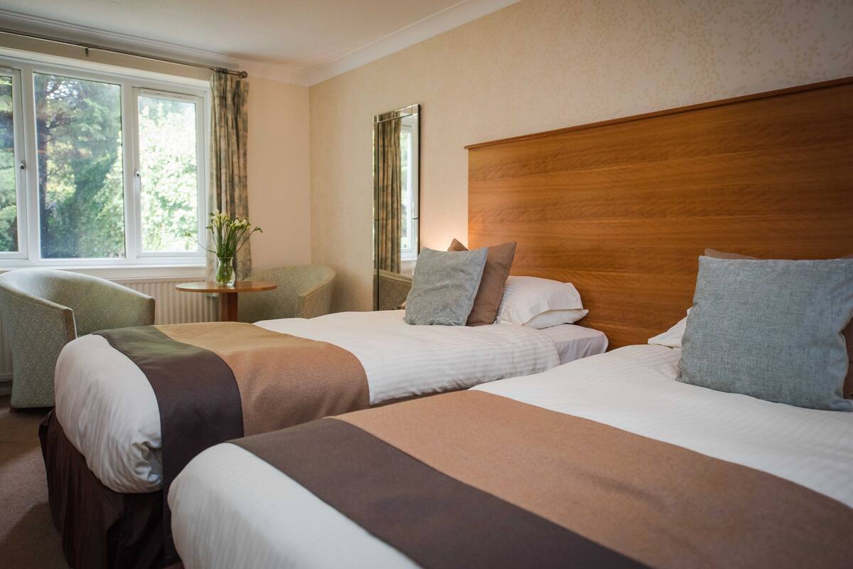 A hotel room with two beds, a sitting area with a chair, and a window with a view of trees.