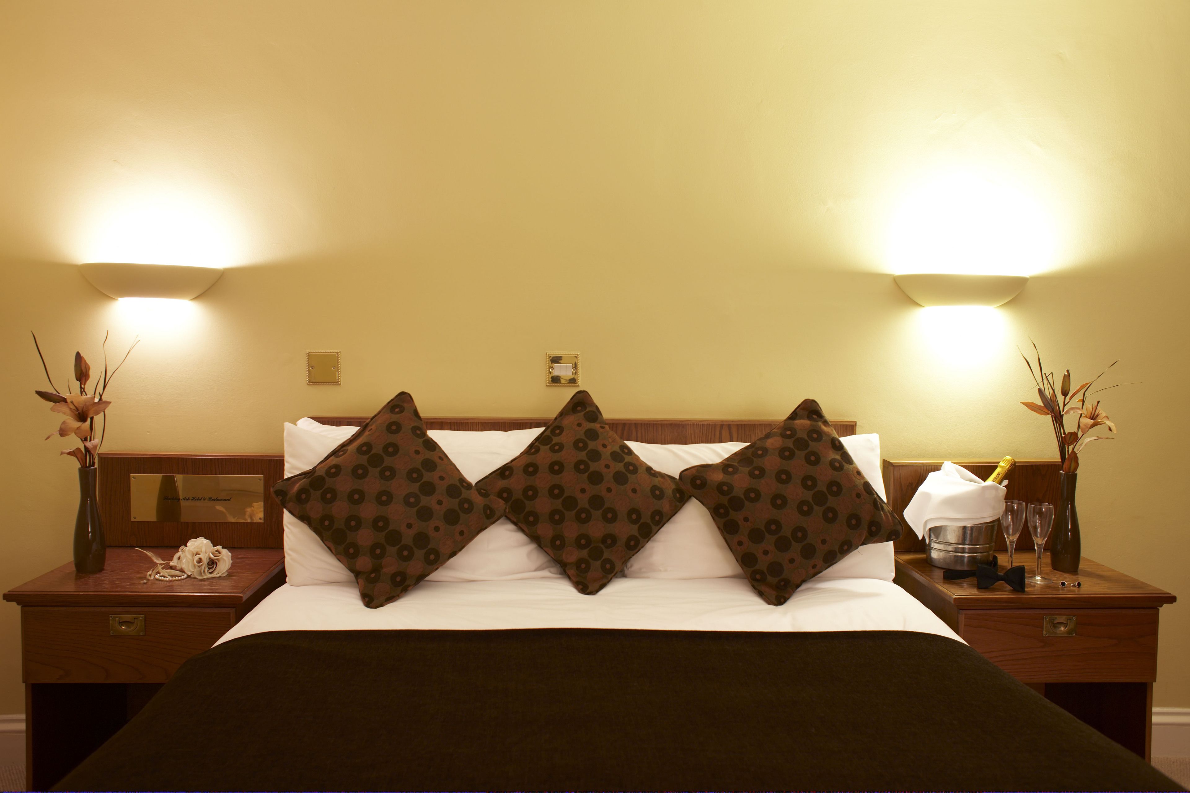 A hotel room with a bed, three patterned pillows, a nightstand with a vase of flowers, and a bottle of champagne.
