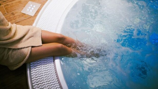 Iemand zit in een hottub, het water borrelt, er is een houten vloer en een bedieningspaneel.