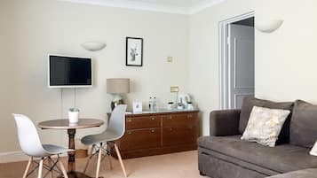 Una sala de estar con un sofá gris, una mesa redonda con dos sillas, un televisor, una lámpara y un aparador.