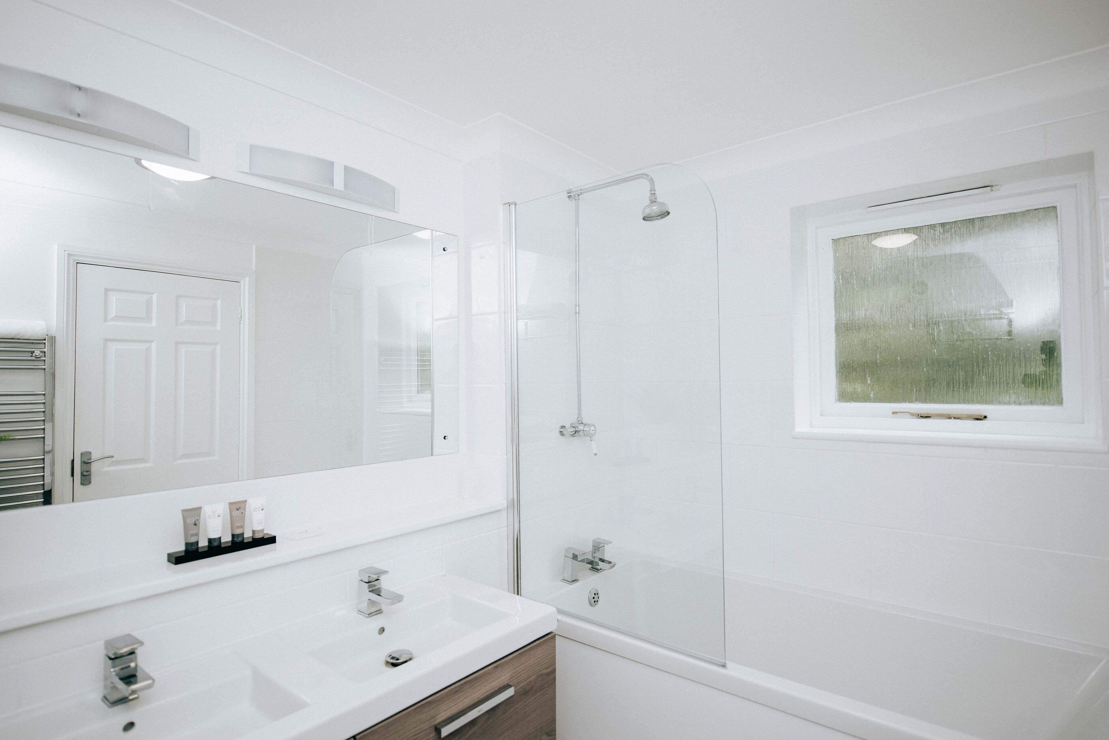 A modern bathroom with a large mirror, a white sink, a shower, and a window.