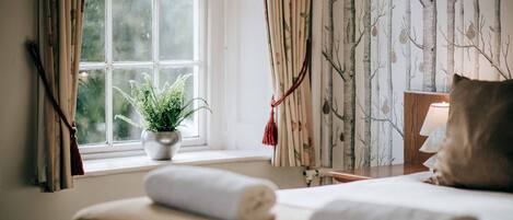 A neatly made bed with a rolled towel, a potted plant by the window, and a lamp on a bedside table.