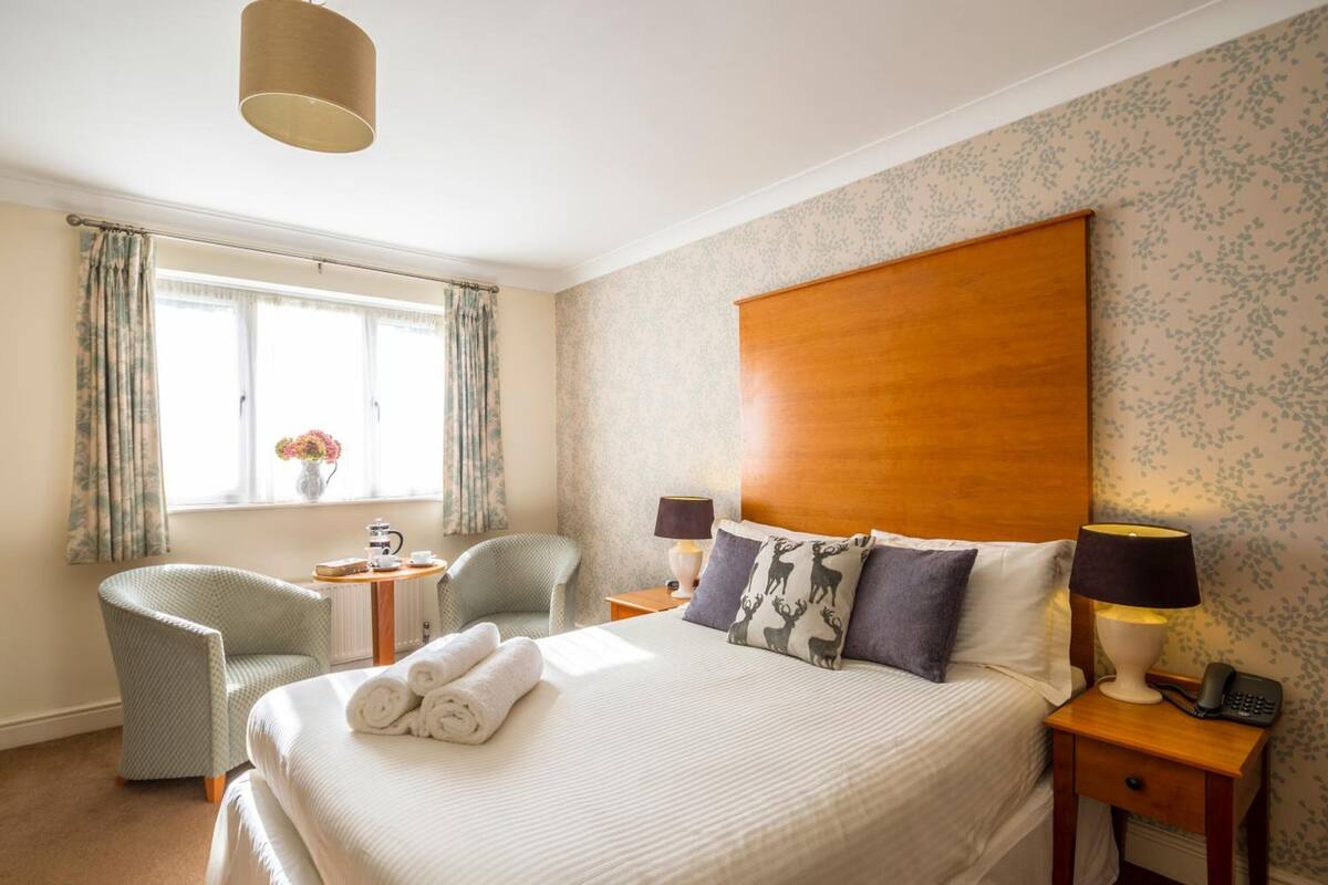 A hotel room with a large bed, two chairs, a small table with a lamp, and a window with curtains.