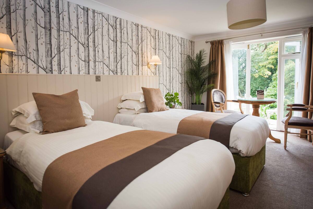 A hotel room with two beds, a large window with curtains, a small table with a chair, and a wall with a tree pattern.